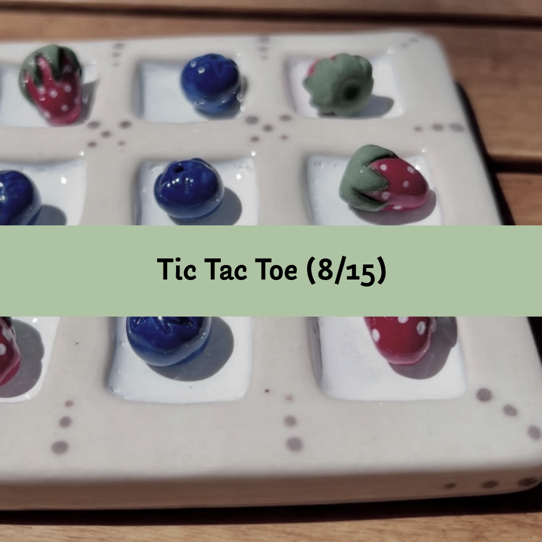 A close-up of a Tic Tac Toe game board with several small glossy beads or tokens placed in the squares, plus a light green text banner across the center reading 'Tic Tac Toe (8/15)'. The board is photographed on a wooden surface.